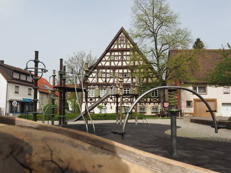 Ein Holzbalken im Vordergrund und dahinter ein Klettergerüst aus Alu-Stangen und Holzelementen mit Rutsche. Im Hintergrund ein Fachwerkhaus: der historische Berchtoldshof sowie grüne Sträucher.