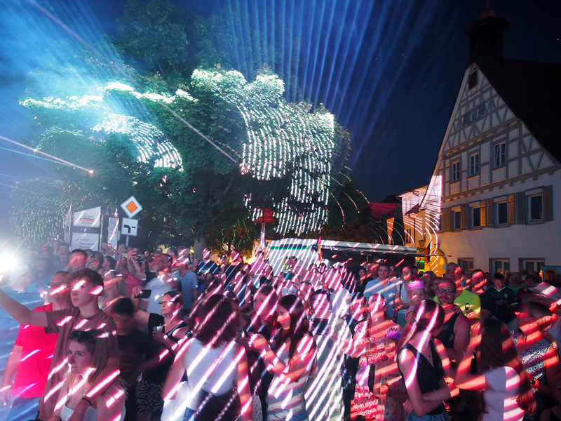 Eine große Menschenmenge steht nachts unter bunten, leuchtenden Laserstrahlen vor einem Fachwerkhaus, dem Uhinger Rathaus.