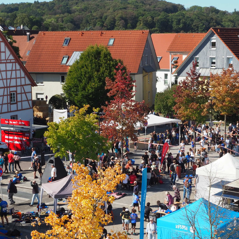 Das Bild zeigt eine geschäftige Straßenszene mit zahlreichen Menschen, die an einem sonnigen Tag zwischen bunten Zelten und Herbstbäumen in einem Dorf mit traditionellen Häusern unterwegs sind.