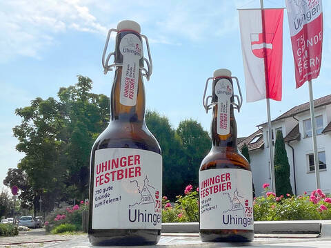 Zwei braune Glasflaschen des Uhinger Festbiers, mit Bügelverschluss, stehen vor dem Rathaus. Im Hintergrund sind die Stadtfahne und Festjahr-Fahne in rot und weiß.
