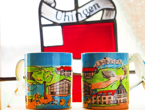 Zwei bunte Kaffee-Tassen stehen auf einem Holzbrett, dahinter ist das rot-weiße Wappen der Stadt in einem Buntglasfenster zu sehen.