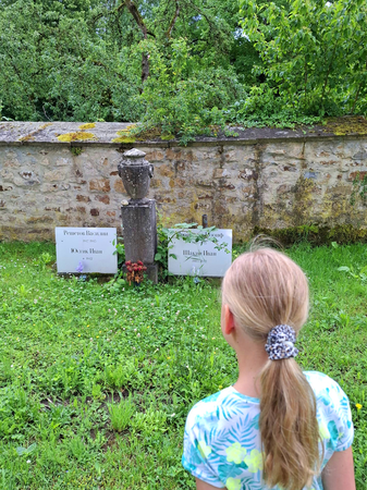 Ein Kind mit blonden Haaren und einem Pferdeschwanz steht vor einer alten Steinmauer und betrachtet Grabsteine auf einem grünen, bewachsenen Friedhof.