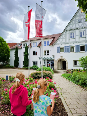 Zwei Mädchen stehen vor einem weißen Gebäude mit Fachwerkfront, halten rote Flyer in den Händen und daneben wehen Fahnen, von denen eine auf das 750-jährige Jubiläum von Uhingen hinweist.