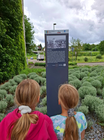 Zwei Kinder stehen vor einer Infotafel mit einer Beschreibung und einem Foto der "Mühle Röhm" in einer grünen Parkanlage.