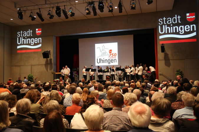 Eine große Menschenmenge sitzt in einem Saal und schaut auf eine Bühne, auf der eine Gruppe von Menschen steht; auf einer Leinwand wird das Jubiläum "750 Jahre Uhingen" angezeigt.