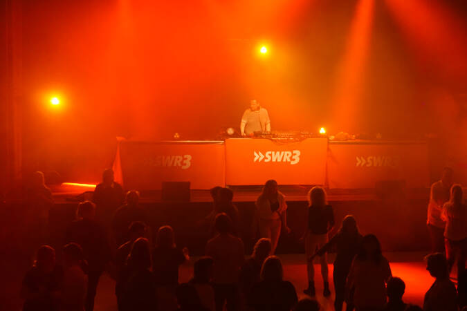 Ein DJ steht auf einer von orangefarbenem Licht beleuchteten Bühne mit SWR3-Logo, während Menschen davor tanzen.