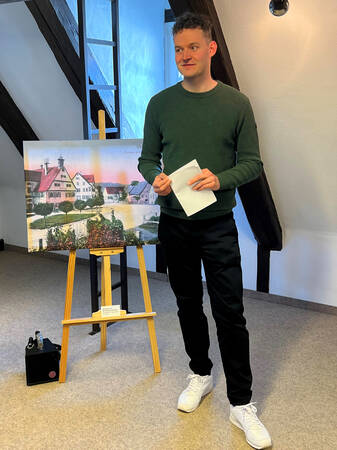 Ein Mann steht lächelnd neben einer Staffelei mit einem Bild einer dörflichen Szenerie aus der Uhinger Historie und hält ein Blatt Papier in der Hand.