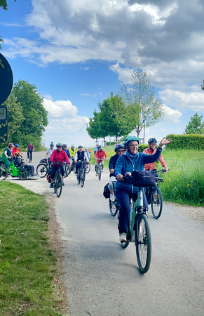 Eine Gruppe von Menschen fährt bei der 750-Jahr-Radtour fröhlich mit Fahrrädern eine ländliche Straße entlang, während der Himmel teils bewölkt ist.