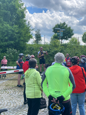 Eine Gruppe von Menschen, viele in Fahrradkleidung, versammelt sich im Freien, während Uhingens Bürgermeister Matthias Wittlinger in gelbem und schwarzem Outfit spricht, während er sich auf einem erhöhten Podest oder einer Plattform befindet, umgeben von Bäumen und einem bewölkten Himmel.