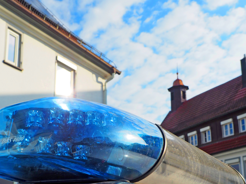 Das Bild zeigt ein blaues Polizeilicht auf einem Fahrzeug im Vordergrund, mit Gebäuden und einem bewölkten Himmel im Hintergrund.