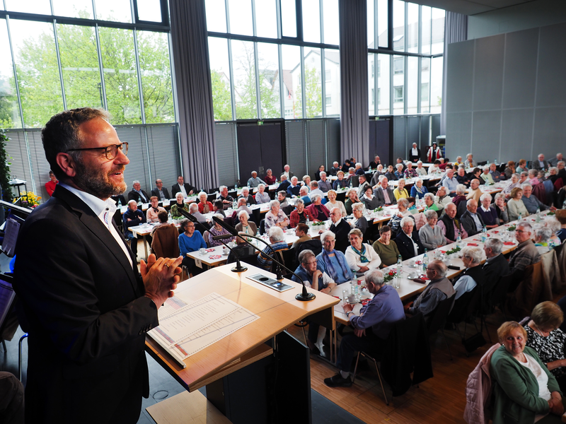 Bürgermeister Matthias Wittlinger hält eine Rede im Uditorium auf der Bühne, vor ihm im Saal sitzen viele Frauen und Männer an Tischen.