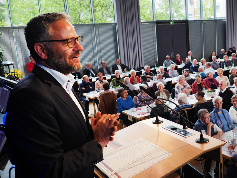 Bürgermeister Matthias Wittlinger hält eine Rede im Uditorium auf der Bühne, vor ihm im Saal sitzen viele Frauen und Männer an Tischen.
