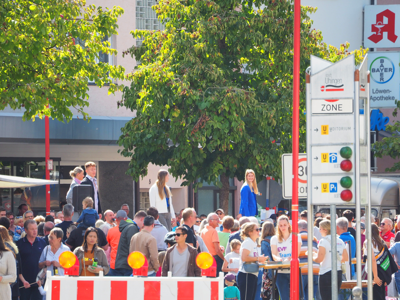 Eine rot-weiße Warnbarke mit orangenen runden Lichtern sperrt eine Straße ab, auf der viele Menschen schlendern. Sie verdecken einen Laufsteg, auf dem Frauen und Männer neue Modetrends präsentieren. Im Hintergrund Bäume und Geschäfte.