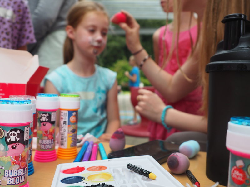 Auf einem Tisch liegt eine Farbpalette, um Gesichter bunt zu schminken. Im Hinergrund wird ein Mädchen geschminkt, andere Kinder und Jugendliche stehen drumherum.