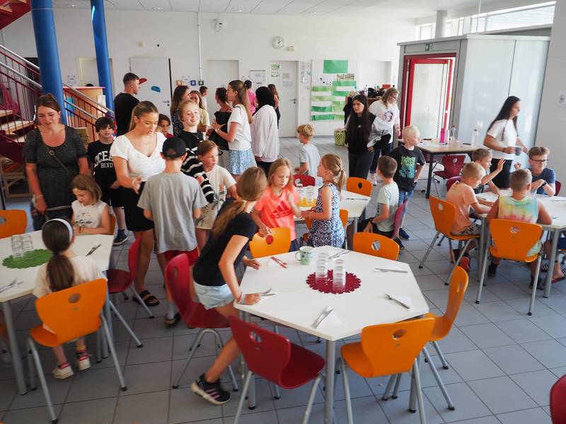 In einer Schulmensa stehen ganz viele Kinder und setzen sich an die gedeckten Tische.