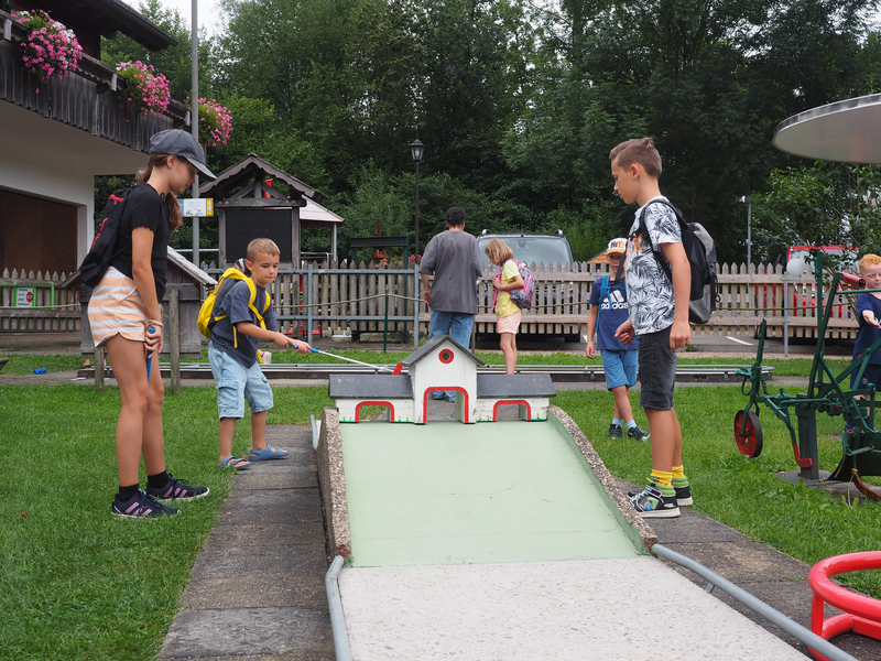 An einer Mini-Golf-Station stehen Kinder mit kleinen Schlägern und ein Junge versucht, den Ball durch ein Haus mit drei Öffnungen hindurch zum Loch zu schlagen.