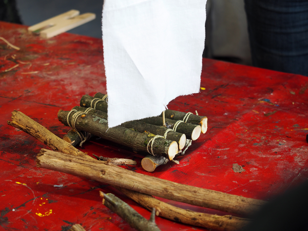 Auf einem Tisch mit roter Wachstischdecke steht ein aus Zweigen und Ästen gebasteltes Floß mit einem kleinen weißen Segel.
