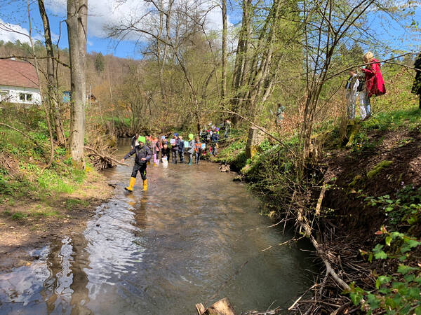 In einem Bach, der Nassach, stehen Kinder mit Gummistiefeln und lassen gebastelte Flöße fahren. Die steilen Ufer der Nassach sind voller Pflanzen.