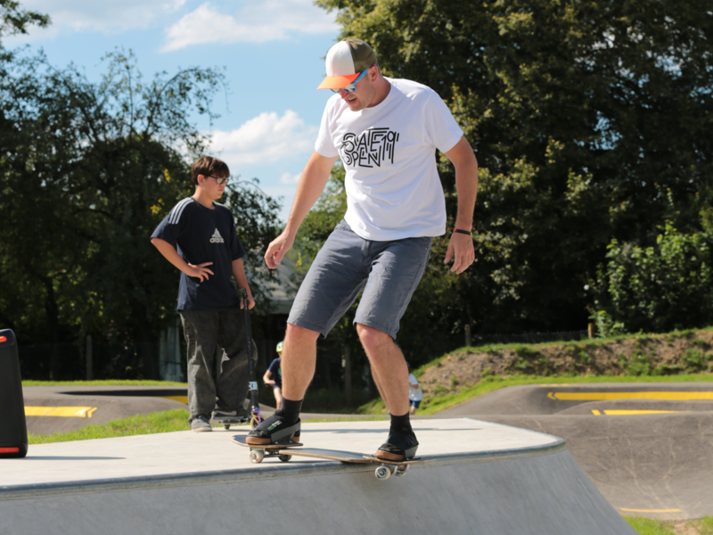 Ein Mann steht in kurzen Hosen auf einem Skateboard an einer Rampe aus grauem Beton und will hinabfahren. Hinter ihm steht ein Junge und hält sein Skateboard.
