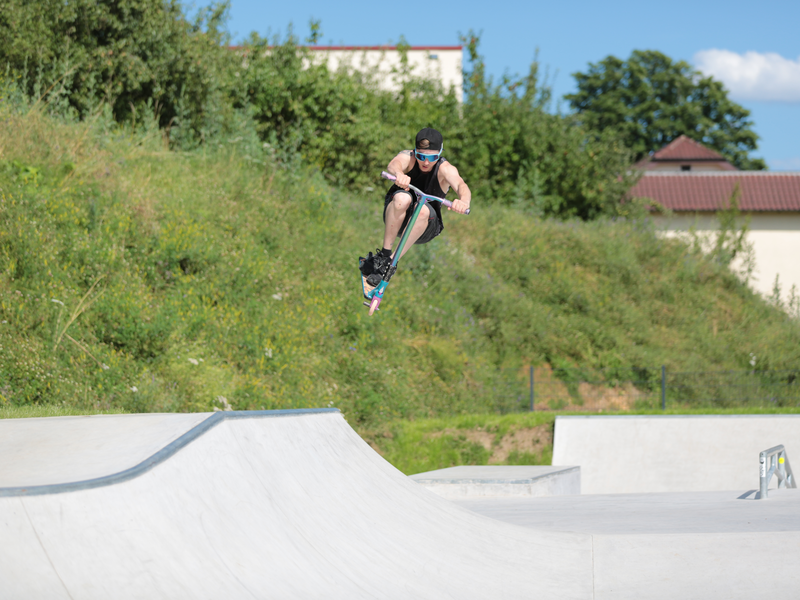 Ein junge fliegt auf einem Roller durch die Luft, unter ihm ist eine Skateanlage mit Betonhindernissen udn im Hintergrund ein mit Gras bewachsender Erdhügel.