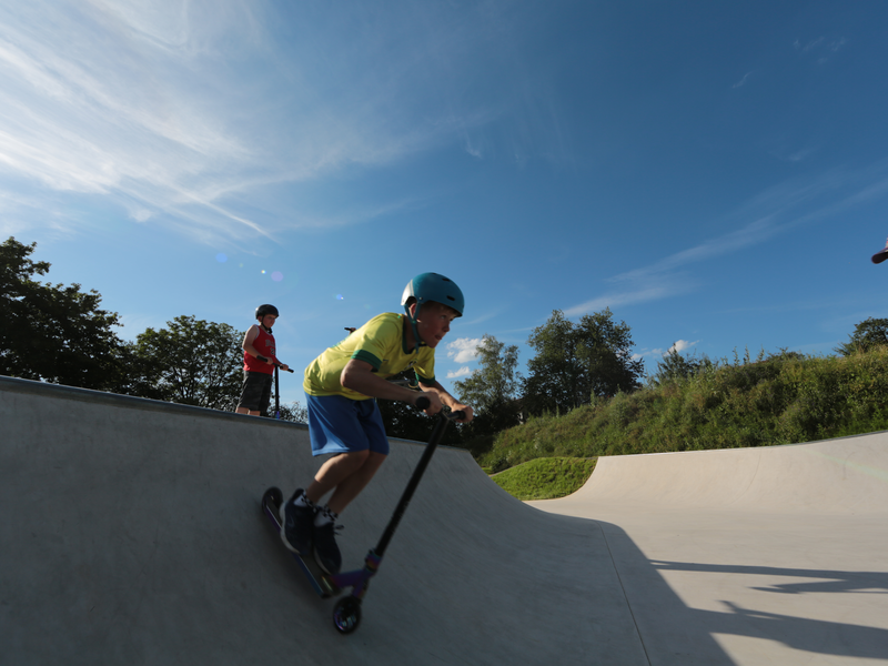 Ein Junge fährt mit einem Roller rasant eine Rampe beim Skatepark hinab, andere schauen ihm zu.