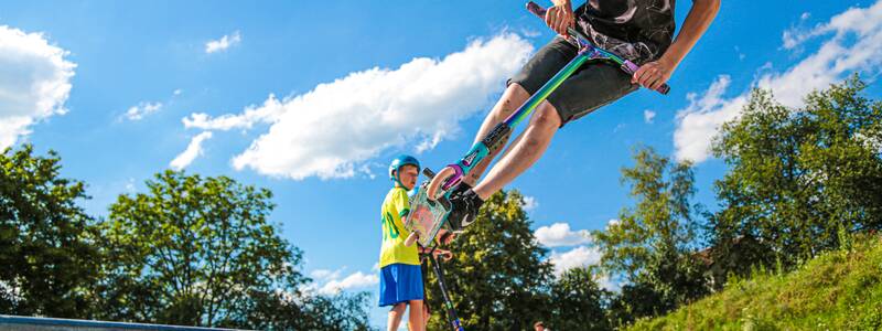 Ein junger Mann springt im Sommer mit einem Roller auf einer Rampe des Skateparks durch die Luft und ein anderer schaut ihm zu.