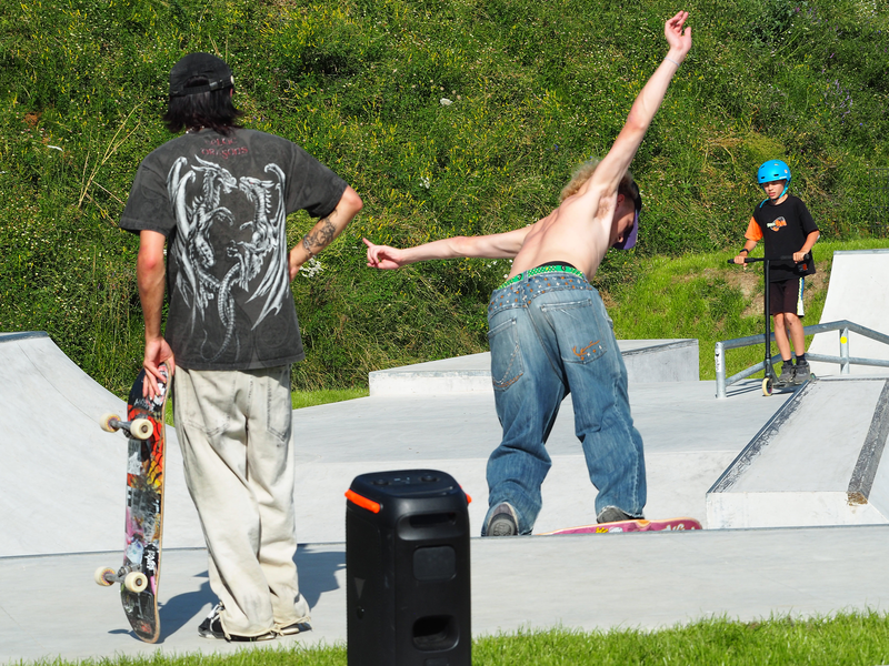 Ein junger Mann fährt mit ausgebreiteten Armen und freiem Oberkörper auf einem Skateboard eine Rampe hinab. Ein anderer hält sein Skateboard in der Hand und schaut zu, im Hintergrund fährt ein Junge auf seinem Roller.