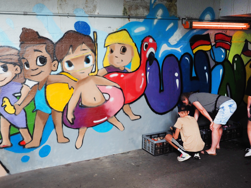 Schüler besprühen für eine Kunstaktion einer Schule die Wand einer Unterführung beim Bahnhof mit Bilder. Auf einem Motiv sind Kinder in Badebekleidung und mit Schwimmringen sowie der angedeutete Schriftzug "Uhingen" mit unterschiedlichen Staatsfahnen.