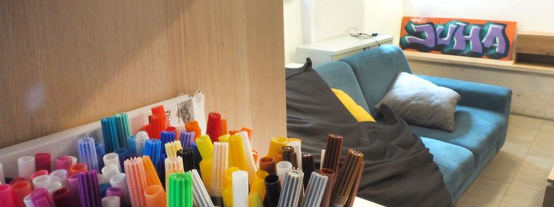 In einem Regal steht eine Kiste mit bunten Filsstiften, dahinter ist ein Sofa mit gelben Kissen und an einer Wand lehnt ein Schild mit der Abkürzung "Juha" für Jugendhaus.