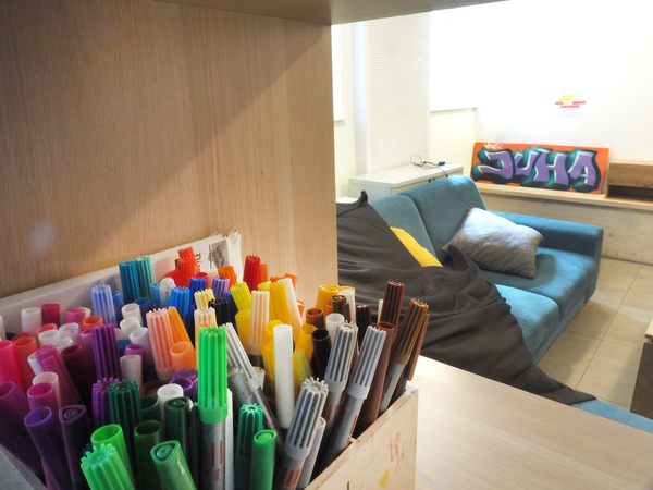 In einem Regal steht eine Kiste mit bunten Filsstiften, dahinter ist ein Sofa mit gelben Kissen und an einer Wand lehnt ein Schild mit der Abkürzung "Juha" für Jugendhaus.