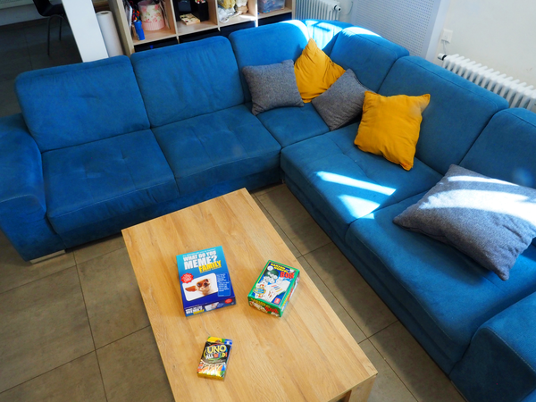 Vor einem blauen Ecksofa mit gelben Kissen steht ein niedriger Couchtisch, auf dem drei Packungen mit Gesellschaftsspielen liegen.