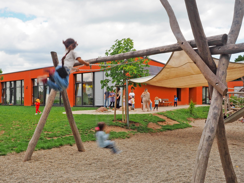 Auf dem Bild schaukeln zwei Kinder auf einem Spielplatz des Kinderhauses Mittlere Mühle neben einem modernen, orangefarbenen Gebäude, während im Hintergrund weitere Personen und Kinder zu sehen sind.