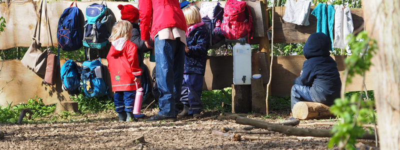 An einem Holzzaun, bestehend aus Pfosten und zwei dicken Brettern, hängen im Wald des Naturkindergartens mehrere Rucksäcke neben- und übereinander. Davor stehen Kinder und eine Erzieherin auf dem mit Holzspänen ausgelegten Waldboden.