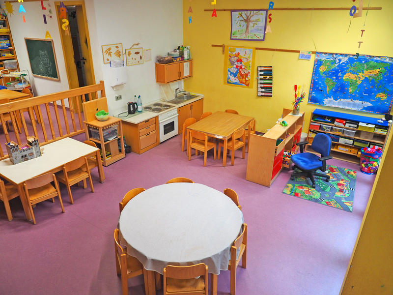 Das Bild zeigt einen hellen, bunt gestalteten Kindergartenraum mit Spiel- und Lernbereichen, mehreren Tischen, Stühlen und einer kleinen Küchenzeile.