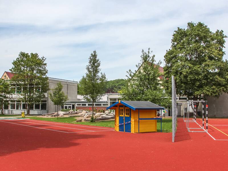 Ein Schulhof mit rotem Sportboden, Laufbahn, buntem Spielhaus, Klettergerüst und umgeben von Bäumen, neben einem Schulgebäude.