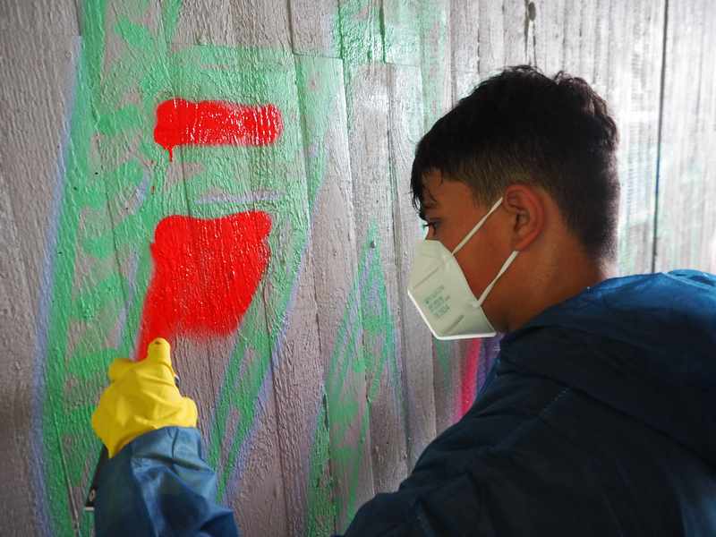 Ein Jugendlicher trägt einen Ganzkörperschutzanzug, hat einen Mundschutz im Gesicht und trägt einen Gummihandschuh und hält eine Spraydose, mit der er auf der legalen Graffiti-Wand vor ihm rote Farbe aufsprüht.