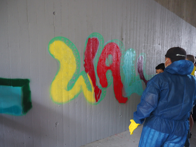 Vor einer grauen Betonwand in einer Unterführung stehen Jugendliche in Ganzkörperschutzanzügen und sprühen auf die legale Graffiti-Wand mit Spraydosen den Schriftzug "Uhingen".