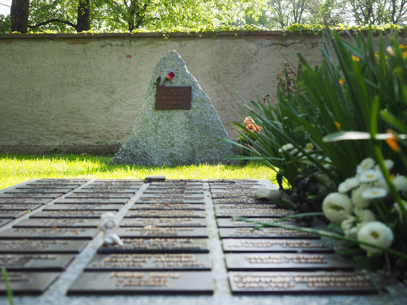Namenstafeln auf dem Boden, im Hintergrund ein mächtiger, spitzer Stein auf einem Rasenfeld vor einer Mauer.