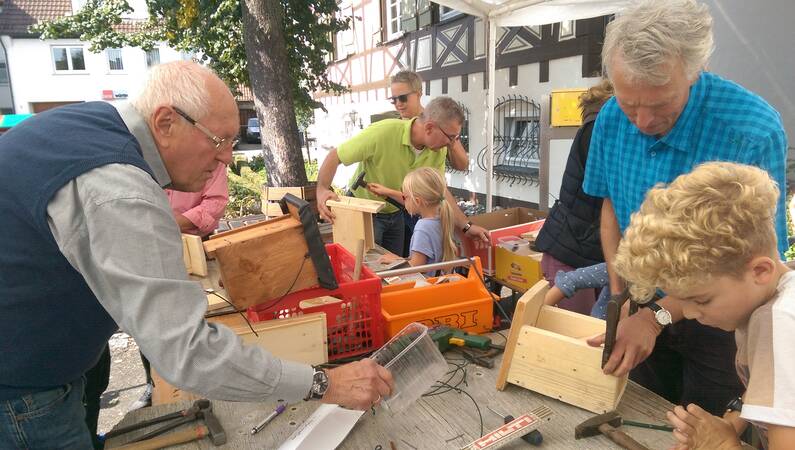 Mehrere Männer stehen mit Kindern in einem Pavillon auf einem Platz. Sie haben Werkzeug und eine Menge Holz auf dem Tisch. Sie zeigen den Kindern, wie man Nistkästen für Vögel baut. 