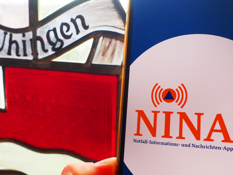 Ein Smartphone zeigt die Benutzeroberfläche der "NINA"-App vor einem Glasfenster mit der Aufschrift "Uhingen".