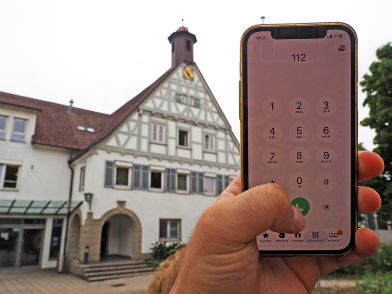 Eine Hand hält ein Smartphone mit der gewählten Nummer 112, während im Hintergrund ein historisch aussehendes Gebäude - das Rathaus der Stadt Uhingen - zu sehen ist.