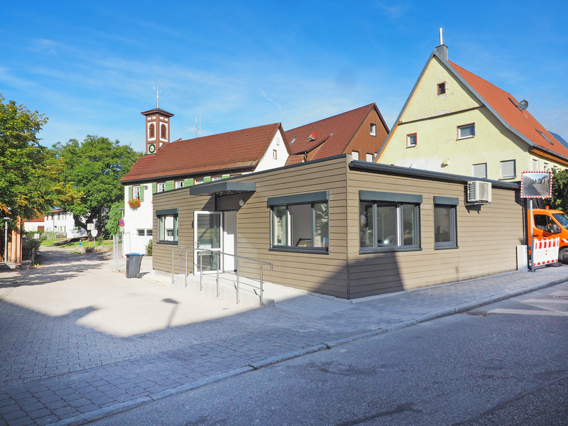 Ein modernes, flaches Gebäude mit großen Fenstern steht in einer sonnigen, gepflasterten Straße neben traditionellen Häusern mit roten Dächern.