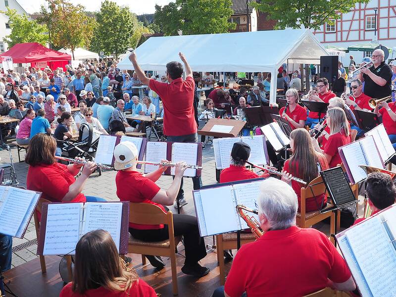 Mitglieder eines Musikvereins sitzen mit Noten und Instrumenten unter freinem Himmel auf einem Auto freien Parkplatz vor vielen Menschen, die auf Bierbänken an langen Biertischen sitzen.  Die Musiker sind von hinten zu sehen, vor ihnen steht der Dirigent mit erhobenen Armen und blickt zum Publikum. Denn er dirigiert nicht nur die Musiker, sondern auch Besucher, die als Massenchor "Freude schöner Götterfunke" singen - begleitet von den Musikern an ihren Instrumenten.