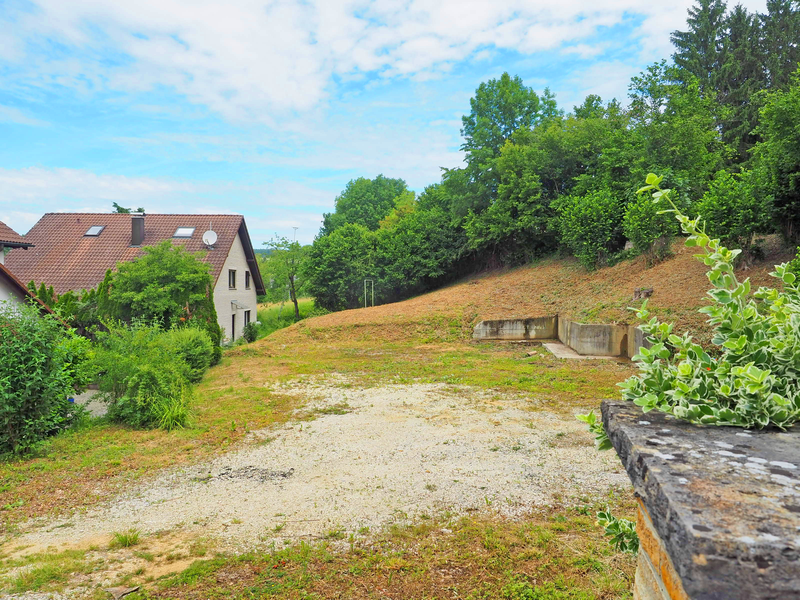 Zwei Häuser mit roten Dächern stehen inmitten von üppiger Vegetation und einem unbebauten, kiesigen Gelände unter einem blauen Himmel. Bei dem Gelände handelt es sich um einen Bauplatz an der Straße "Am Giebel", den die Stadt Uhingen verkauft.