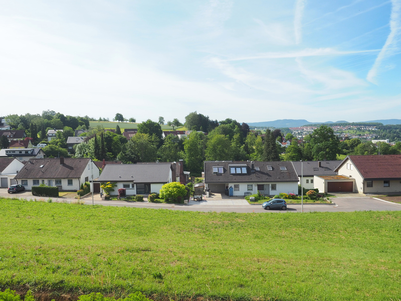 Das Baugebiet Bruckstraße im Uhinger Stadtteil Holzhausen: Eine idyllische ländliche Szene mit mehreren Einfamilienhäusern, einer grünen Wiese im Vordergrund und einem bewaldeten Hügel im Hintergrund unter einem blauen Himmel.