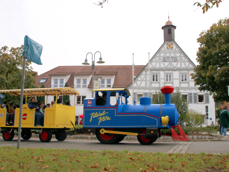 Ein Mini-Zug fährt auf einer Straße und zieht zwei Waggons, in denen Menschen am mehrgeschossigen Rathaus von Uhingen vorbeifahren.