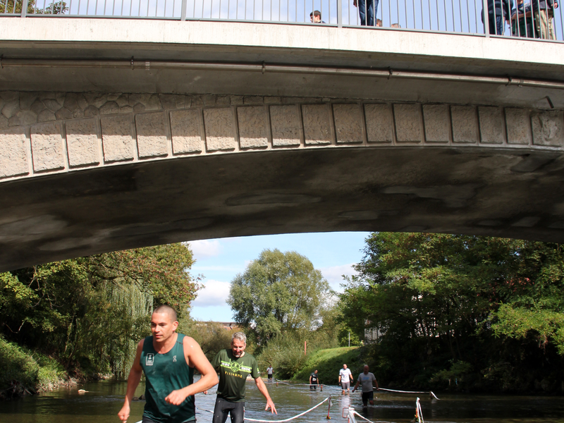 Zwei Männer rennen durch das knöchelhohe Wasser des Flusses Fils, auf einer über ihnen befindlichen Brücke schauen Passanten hinunter.