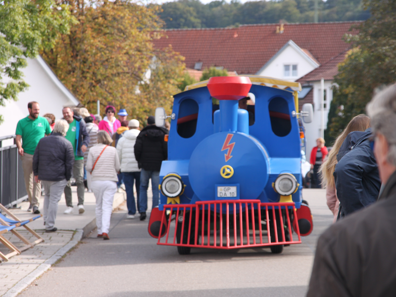 Ein blauer für die Straße als Zug umgebauter Traktor, der Filstalblitz, fährt über eine Brücke an Passanten vorbei.