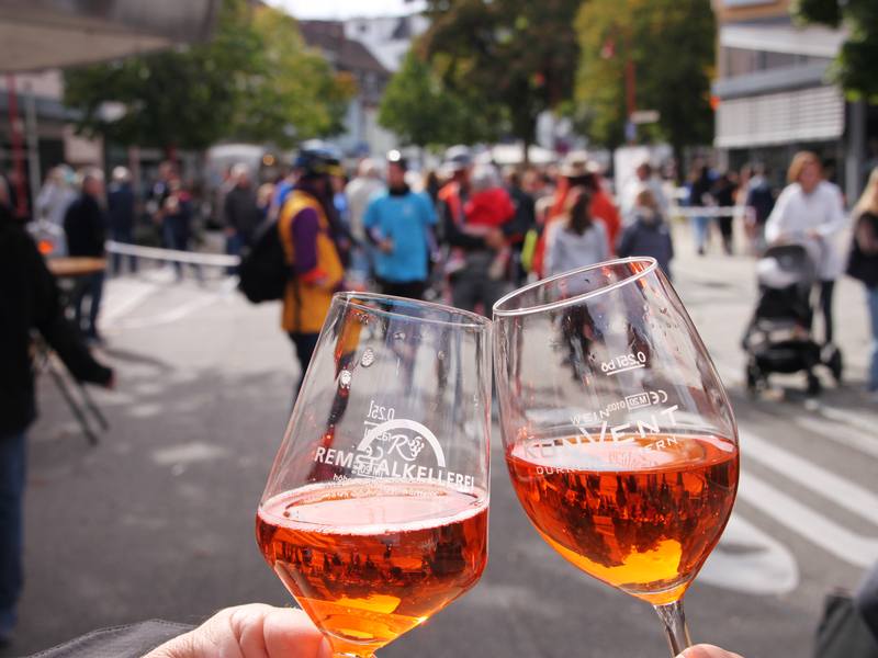 Zwei Weingläser mit orangener Flüssigkeit stoßen aneinander, im Hintergrund flanieren Menschen über eine Straße.
