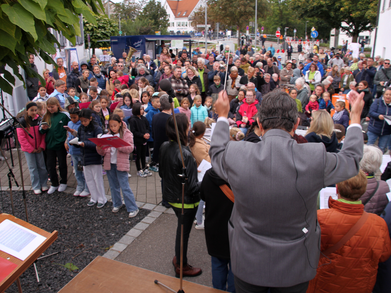 Ein Dirigent wedelt mit dem Taktstock, vor ihm singt eine große Menschenmenge.